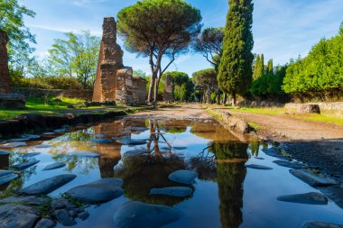 Appia Antica, Roma, bir sonbahar gününde mavi gökyüzü ve bulutlarla gelen fırtınadan sonra su birikintisindeki mezarların kalıntılarını yansıtır. Regina Viarum, İtalya.