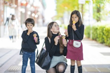 Anne ve iki çocuk dışarıda gülümseyerek dondurma yiyorlar.