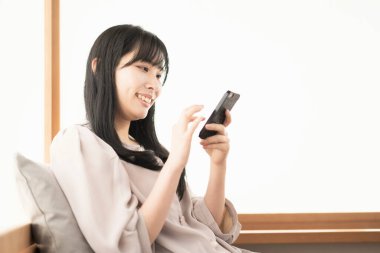 Asyalı kadın evde oturuyor ve akıllı bir telefon kullanıyor.