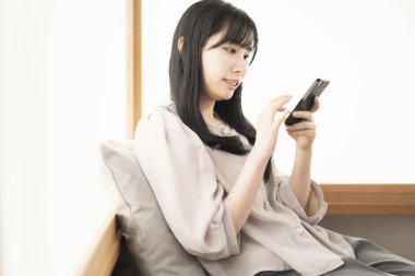 Asyalı kadın evde oturuyor ve akıllı bir telefon kullanıyor.