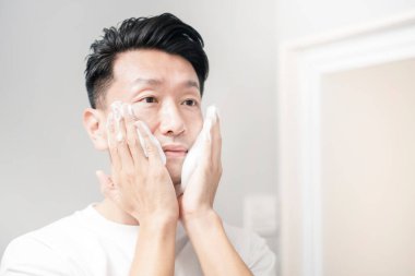 Orta yaşlı bir adam yüzünü lavaboda köpükle yıkıyor.