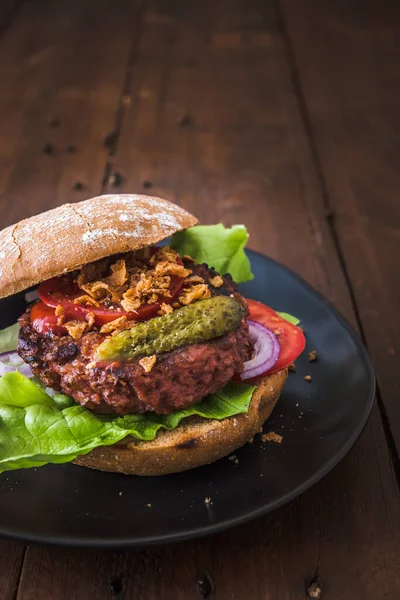 Burger with vegetarian hamburger patty, salad, tomatoes and roasted onions on a black plate on wooden table, vertical stock photo with copy space