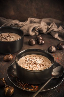 Parmesanlı kestane çorbası, kırmızı biber taneleri ve koyu kahverengi arka planda siyah bir kasede iplikler.