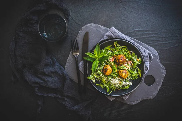 Roket salatalı siyah kase, domatesli kavrulmuş çam tohumu, parmesan peyniri ve koyu arka planda kızarmış vejetaryen topları.