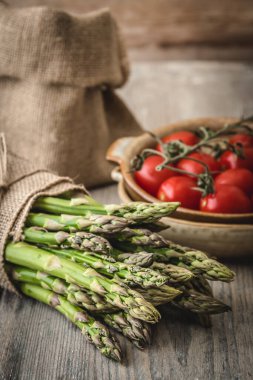Kırsal ahşap bir masada yeşil kuşkonmaz, arka planda domates ve jüt çuvalı, dikey