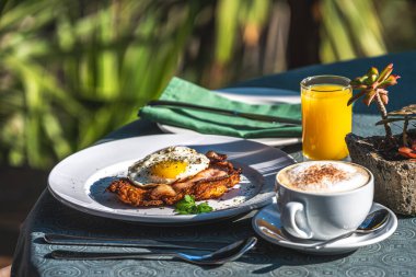 Güneşte kahvaltı, kapuçino, patates rosti, kızarmış yumurta, pastırma ve portakal suyu, yeşil arka planda.