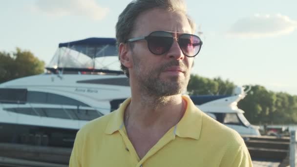 Przystojny, dojrzały mężczyzna w okularach przeciwsłonecznych, przystań jachtowa w marinie w tle. — Wideo stockowe