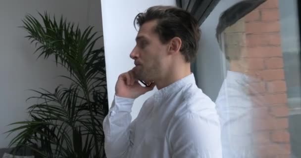 Young businessman answers the phone. Man talking on mobile phone with client. Young business entrepreneur in casuals standing by a window and talking over smartphone in office. — Stock Video