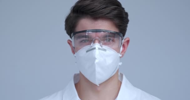 Portret van een knappe arts of wetenschapper met een witte jas en masker, en een veiligheidsbril die naar de camera kijkt, geïsoleerd op een witte achtergrond. — Stockvideo