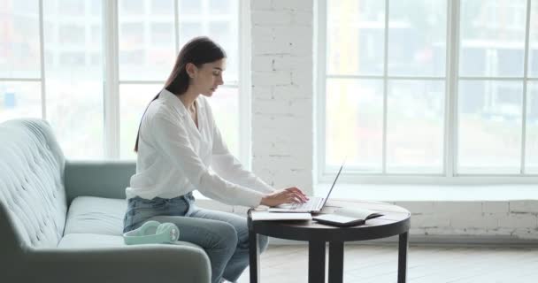 Przemyślana młoda bizneswoman brunetka pisząc na laptopie siedzi sofa. — Wideo stockowe