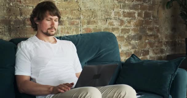 Фрілансер працює дистанційно з дому на ноутбуці, сидячи на дивані , — стокове відео