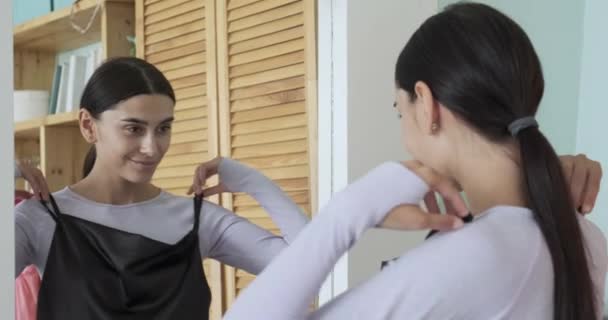 Молодая улыбающаяся брюнетка проверяет черное коктейльное платье в гардеробе, отражающееся в зеркале дома.. — стоковое видео