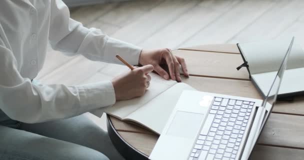 Vista de cerca de la joven mujer escribiendo en bloc de notas y mirando a la computadora portátil con tutorial. — Vídeos de Stock