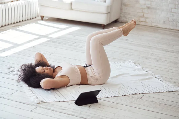 Zwarte jonge vrouw kijken video tutorial op tablet strekt benen en armen thuis met grote ramen in de ochtend. — Stockfoto