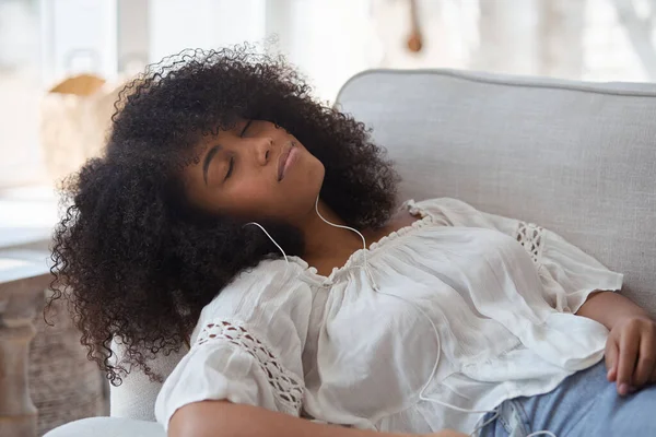 Una joven y relajada mujer negra sonriente con auriculares, descansando en un cómodo sofá —  Fotos de Stock