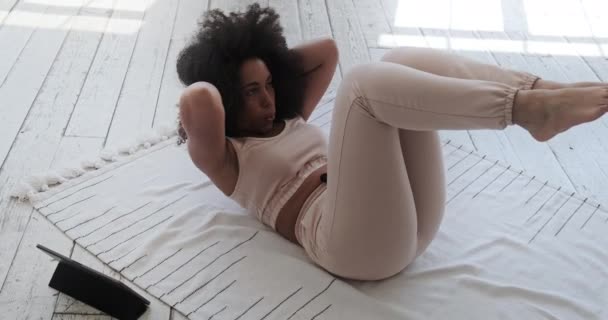 Jeune femme noire regardant tutoriel vidéo sur tablette étire les jambes et les bras à la maison avec de grandes fenêtres le matin. — Video