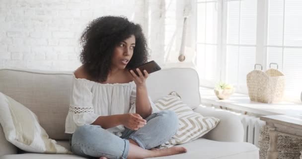 Młoda uśmiechnięta birasowa kobieta nagrywa wiadomość audio do znajomego za pomocą smartfona siedzi na szarej kanapie w domu. — Wideo stockowe