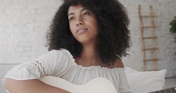 Молодая афро-американка расслабляется с музыкой и играет на акустической гитаре дома. — стоковое видео