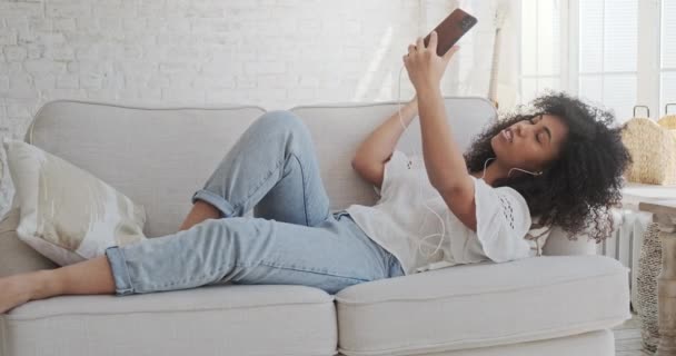 Mulher negra feliz em fones de ouvido olhando para o smartphone, desfrutando de reunião de vídeo com amigos e colegas — Vídeo de Stock