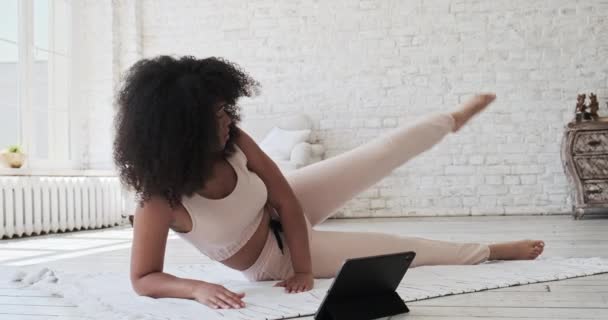 Атлетичная молодая черная женщина смотрит видео-учебник на планшете, делая упражнения дома с большими окнами по утрам. Смотреть — стоковое видео
