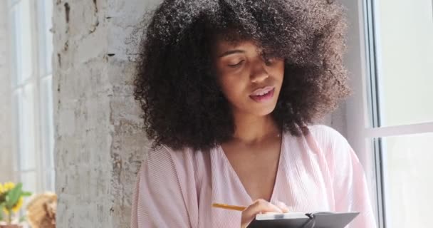 Задумчивая черная молодая женщина с вьющимися волосами оглядывается и пишет в бумажной тетрадке у окна — стоковое видео