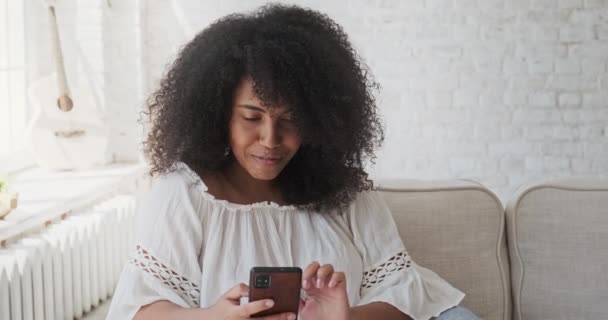 Fiatal fekete mosolygós nő felvétel audio üzenet barátjának segítségével okostelefon ül szürke kanapén — Stock videók