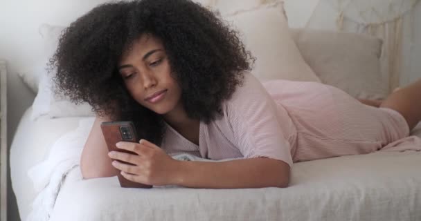 Una mujer negra relajada y tranquila se acuesta en su cama y disfruta de las redes sociales en su teléfono inteligente — Vídeo de stock