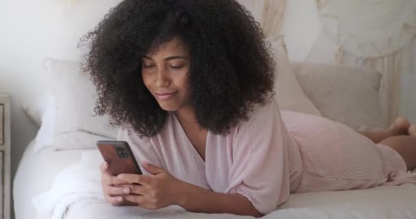 En frissigt hårig, snygg afro amerikansk kvinna ligger på en säng med en smartphone — Stockvideo