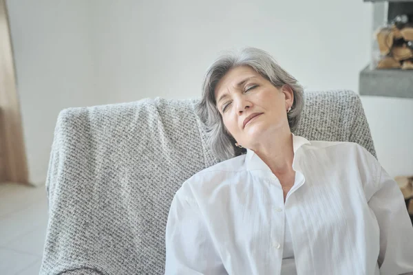 Descansando sonriente anciana sentada en un cómodo sofá estirándose, calmada, meditando — Foto de Stock