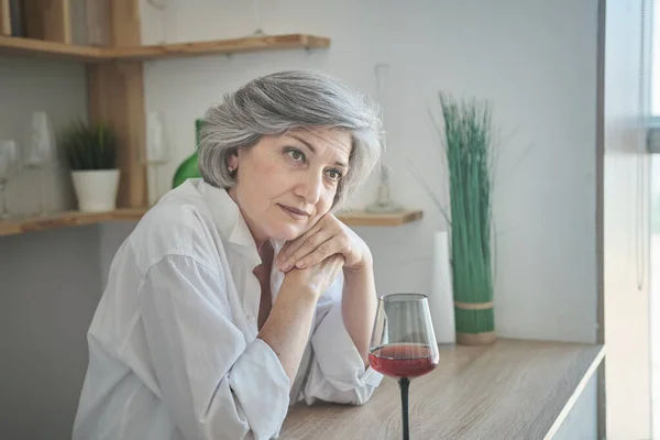 Portret van een vrolijke oudere vrouw met een glas wijn in een licht comfortabel appartement — Stockfoto