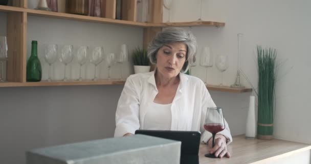 Ältere 60-jährige Frau steht mit Tablette, trinkt Wein und genießt Videoanruf — Stockvideo