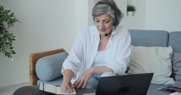 Старшая взрослая женщина в наушниках принимает участие в бизнес-видеоконференции — стоковое видео