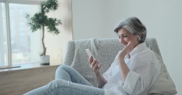 Счастливая пожилая женщина в наушниках смотрит на мобильный экран, наслаждаясь видеовстречей — стоковое видео