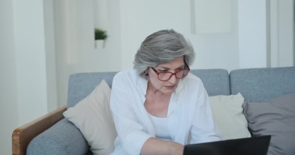 Bedachtzame oudere vrouw freelancer dragen bril typen op laptop zitbank. — Stockvideo