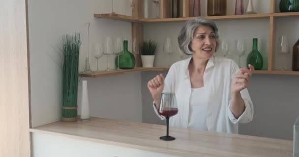 Uma mulher mais velha dança na cozinha enquanto bebe vinho e se sente livre de anos seniores. — Vídeo de Stock