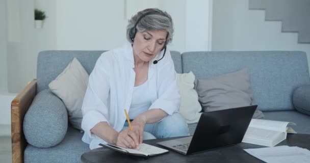 Senior volwassen oude vrouw werken op afstand van modern huis via video conferentie, het maken van notities over het project — Stockvideo