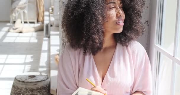 Задумчивая молодая черная женщина с вьющимися волосами смотрит вокруг и пишет в бумажной записной книжке у окна. — стоковое видео