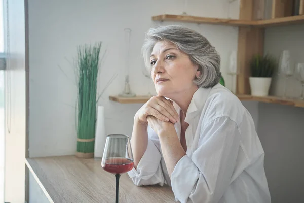Portret van een vrolijke oudere vrouw met een glas wijn in een licht comfortabel appartement — Stockfoto