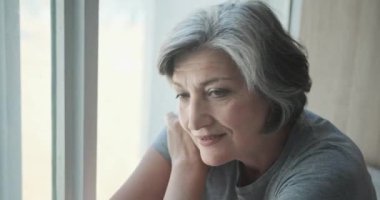 Rehabilitasyon merkezinde düşünceli yaşlı bir kadın pencerenin kenarında oturuyor.