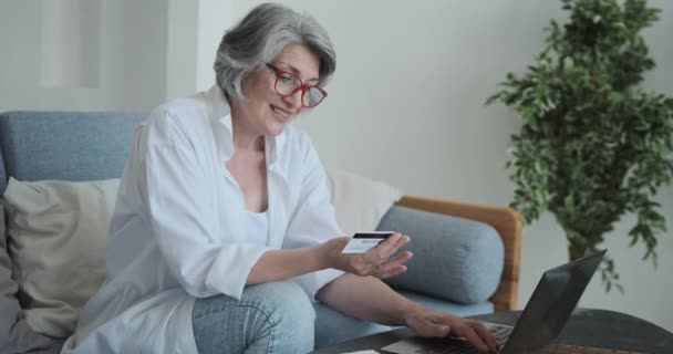 Sonriente cliente de banco mujer anciana haciendo pagos rápidos en línea con su tarjeta de crédito y portátil — Vídeo de stock