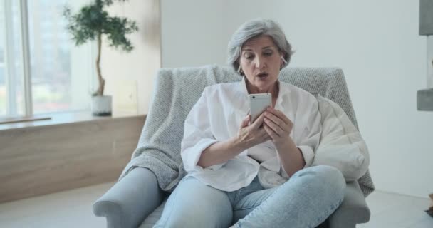 Fröhliche ältere Frau mit Ohrhörern blickt in ihre Smartphone-Kamera und genießt einen Videoanruf — Stockvideo