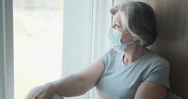 Ältere depressive Frau mit Maske sitzt am Fenster und hofft auf ein Ende der Epidemie und Freiheit. — Stockvideo