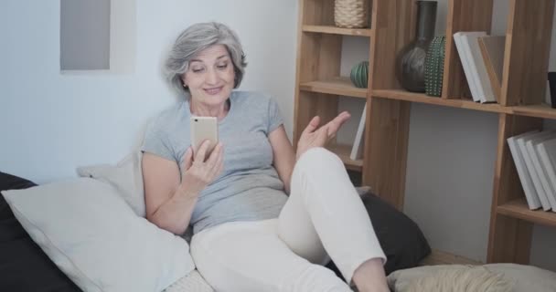 Веселая пожилая женщина смотрит на камеру своего смартфона, наслаждаясь видеозвонком — стоковое видео