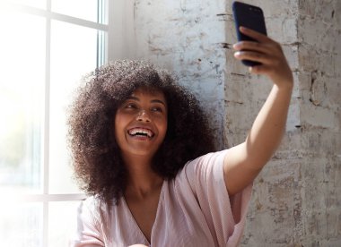 Genç melez kadın akıllı telefon ekranına bakıyor, selfie çekiyor.