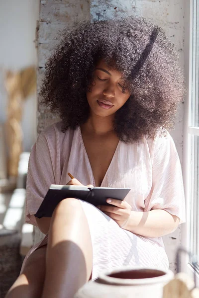 Una joven negra pensativa con el pelo rizado mira a su alrededor y escribe en un cuaderno de papel cerca de la ventana —  Fotos de Stock