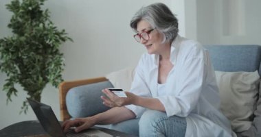 İnternetten almak için kredi kartı ve dizüstü bilgisayar kullanan beyaz, yaşlı bir kadın..