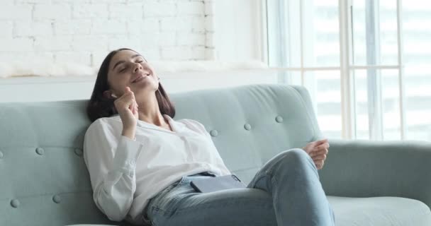 Молодая женщина отдыхает на удобном диване, наслаждаясь прослушиванием лаундж-музыки в прямом эфире через приложение смартфона — стоковое видео