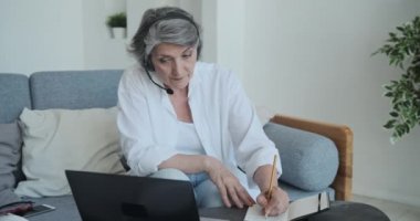 Akıllı ve motive olmuş yaşlı kadın internette yeni beceriler öğrenmek için dizüstü bilgisayar ve kulaklık kullanıyor.