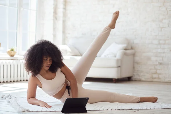 Atletische jonge zwarte vrouw kijken video tutorial op tablet doen oefeningen thuis met grote ramen in de ochtend. Kijk. — Stockfoto