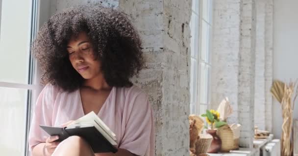 Nachdenkliche schwarze junge Frau mit lockigem Haar schaut sich um und schreibt in ein Notizbuch — Stockvideo